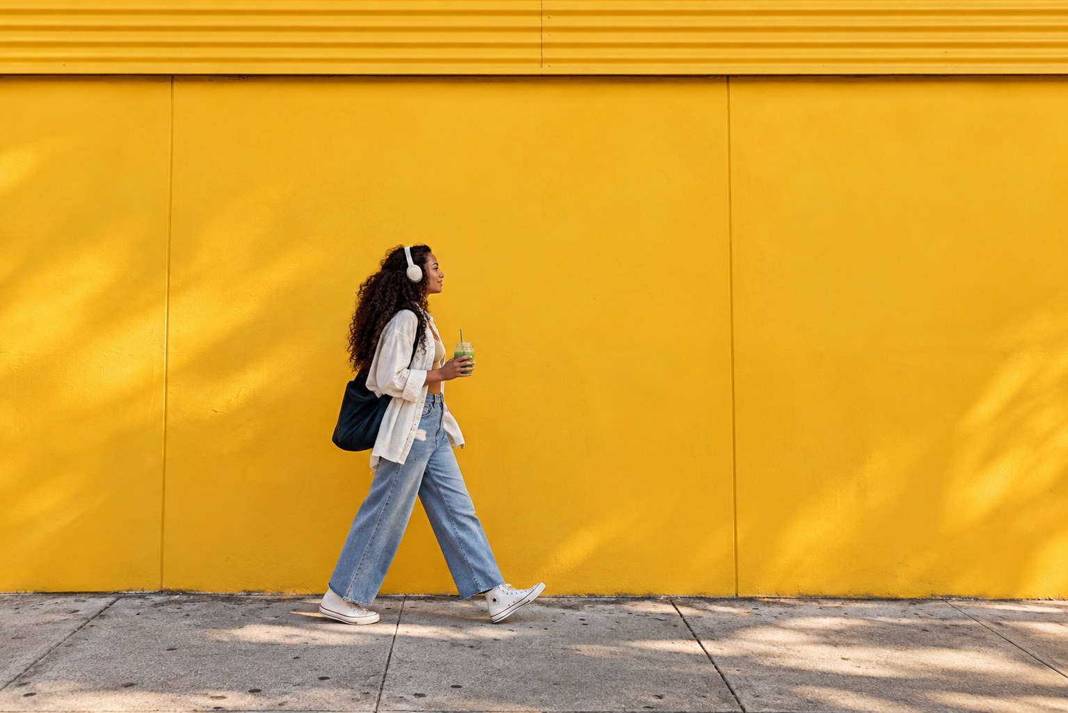 A woman walking past a bright yellow wall with headphones and a green juice