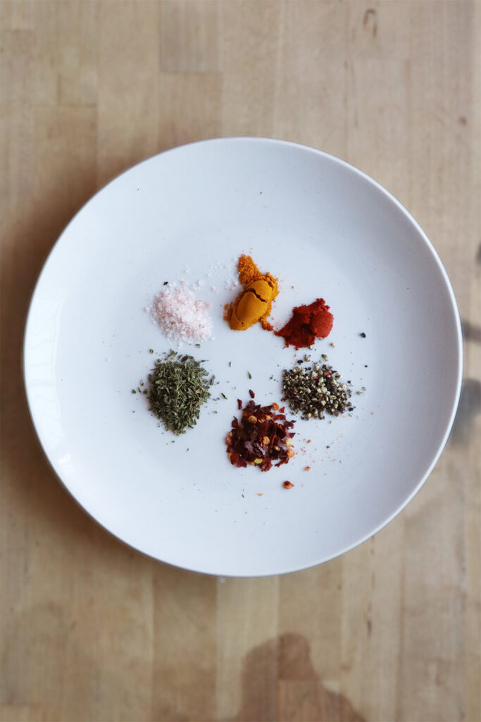 Spices on a white plate. Salt, turmeric, paprika, rainbow pepper, red chili pepper flakes, and oregano. All added to scrambled tofu.